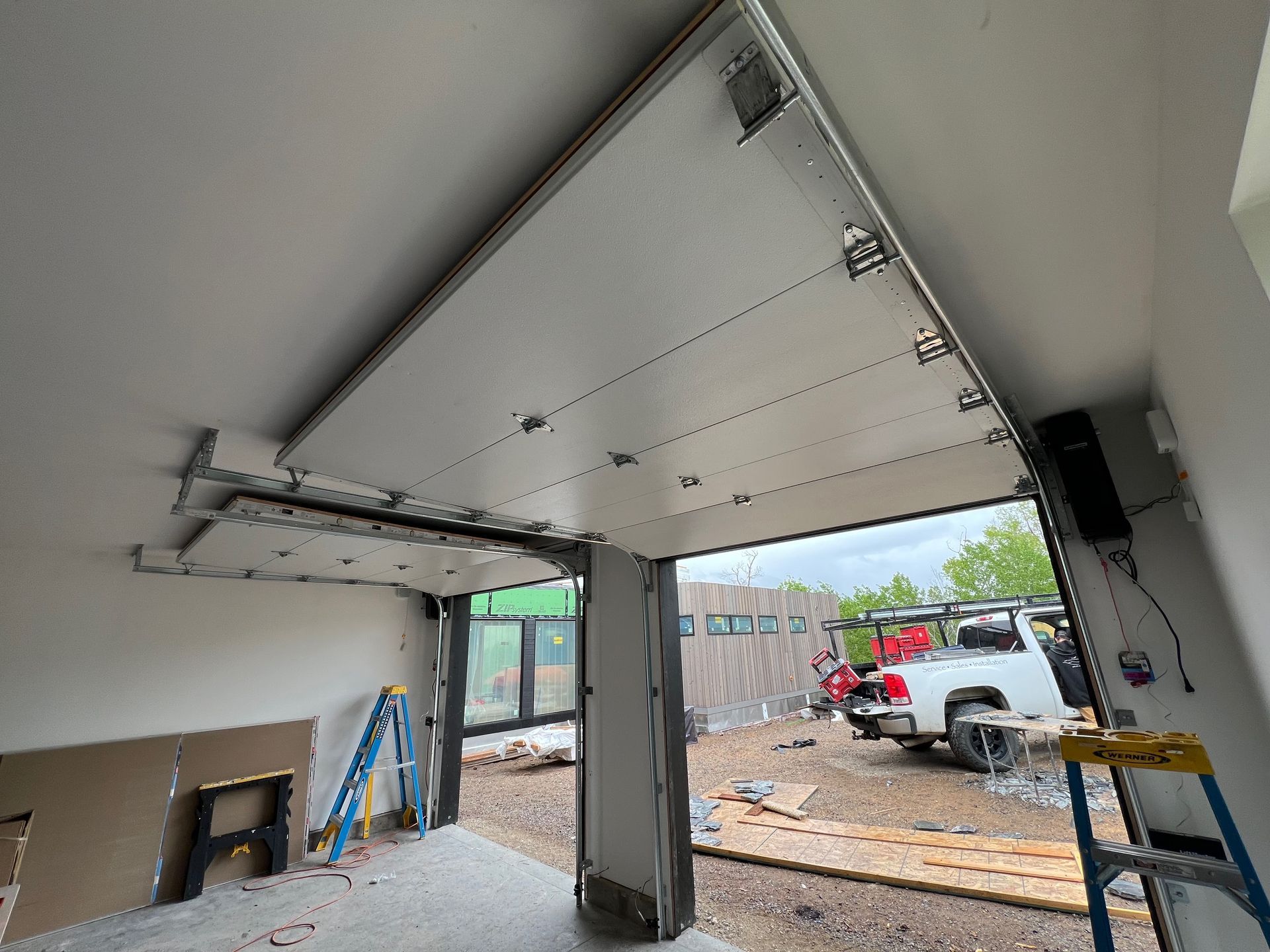 Garage ceiling with installed white panels, track, and cables. An open doorway reveals construction outside, including a truck and building.