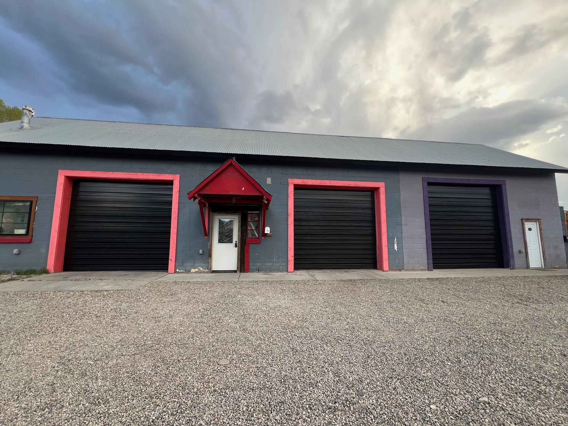 A gray building with three black garage doors framed in pink, purple, and red. A white door with a red awning is in the center, with a gravel parking lot in the foreground.