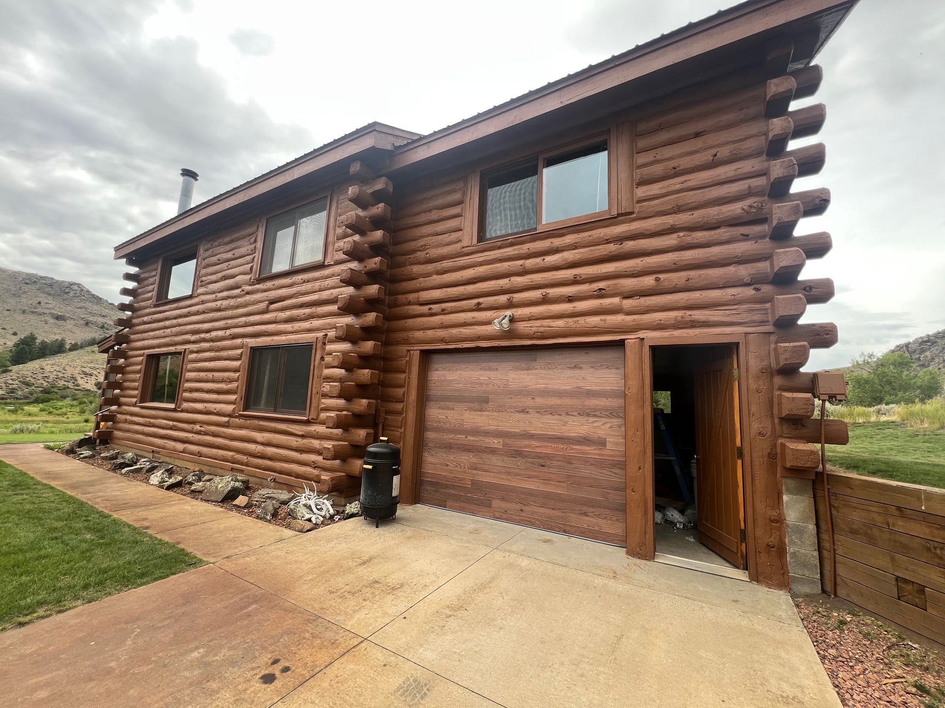 Two-story brown log cabin with a garage door open, parked in a concrete driveway, against a cloudy sky.