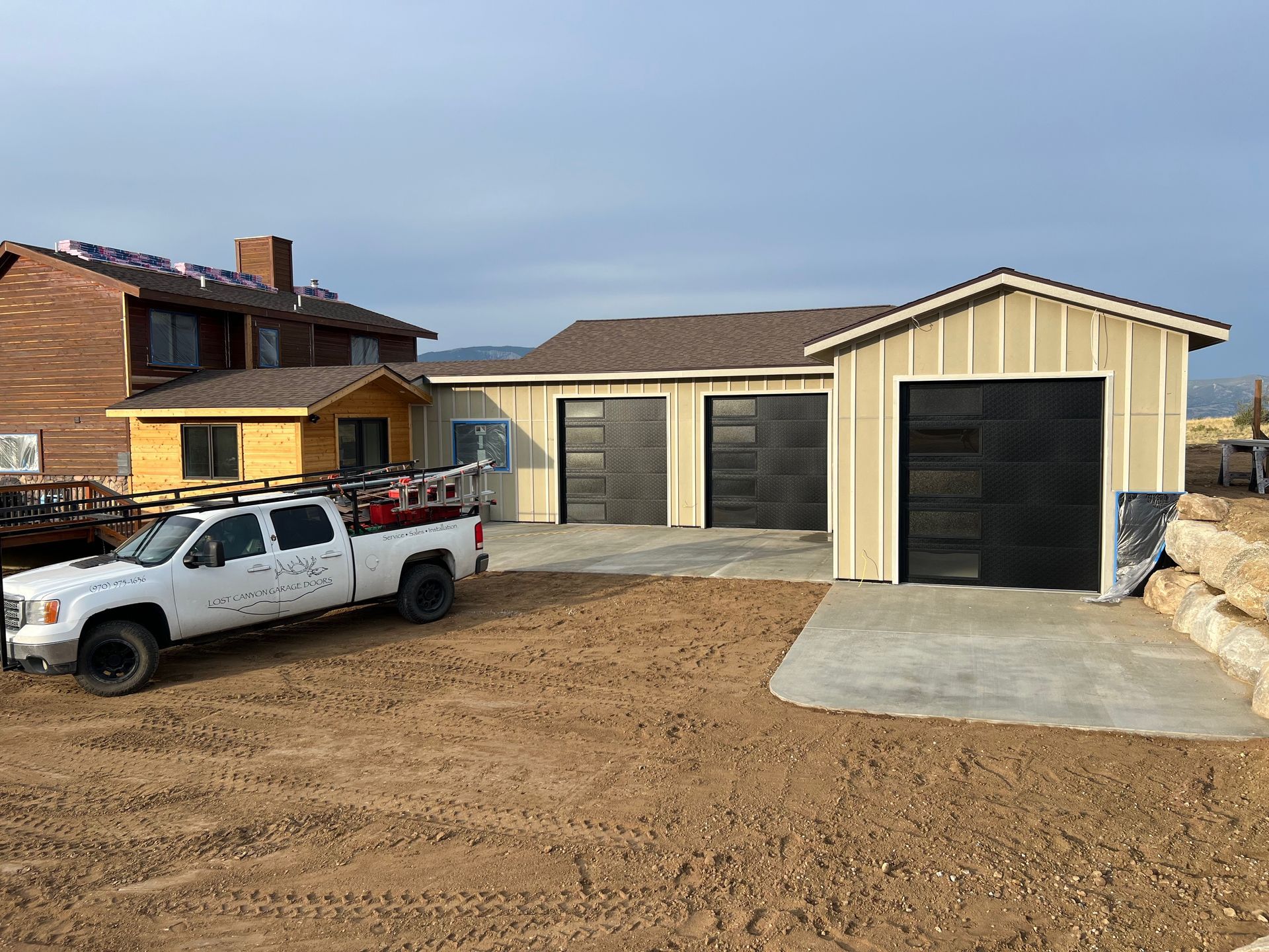A white pickup truck parked in a dirt lot in front of a newly constructed garage with black garage doors.