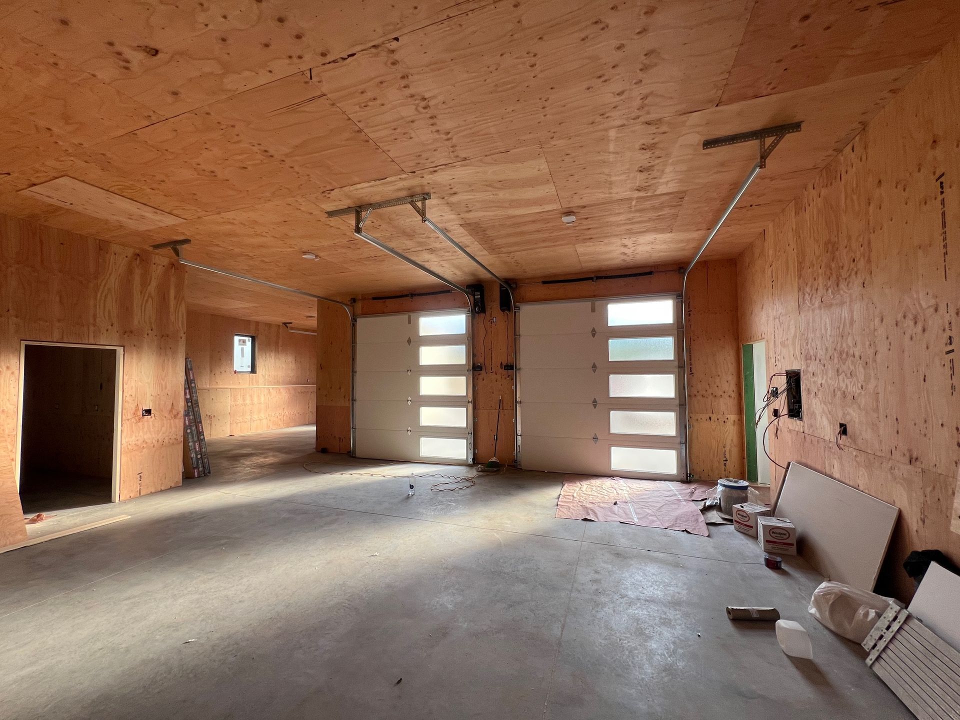 Interior view of a garage under construction, with wood paneling, concrete floor, two garage doors, and tools scattered around.