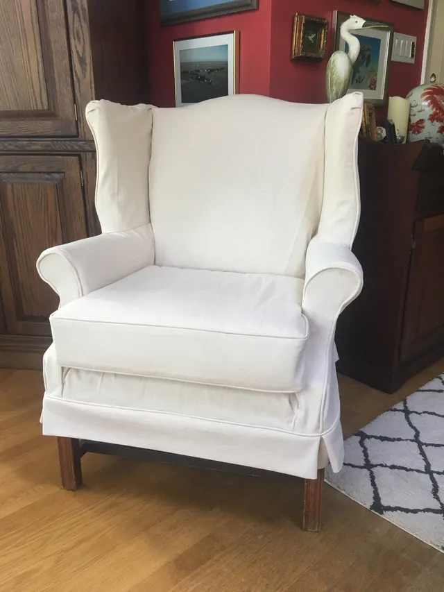 White upholstered wingback chair on hardwood floor, against a red wall with artwork.