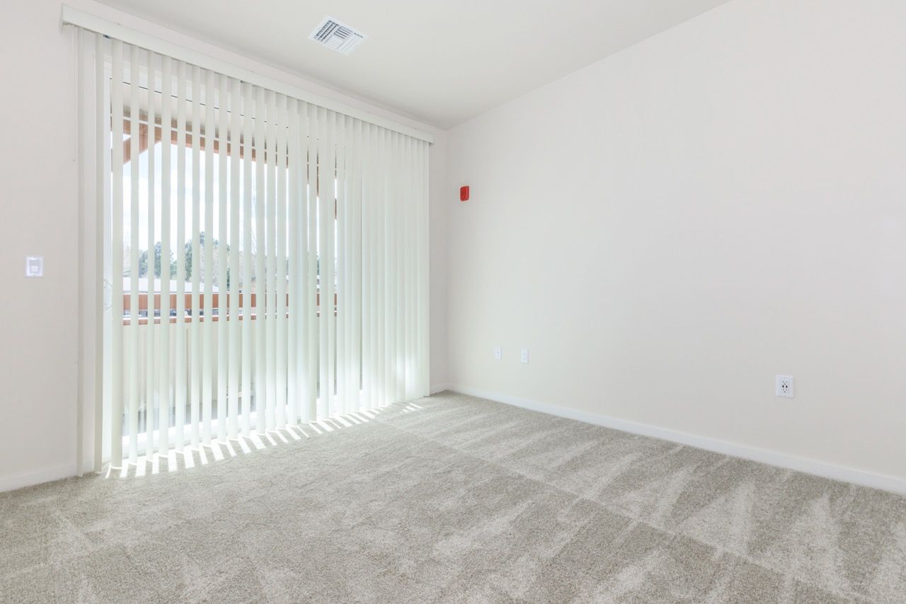 Empty beige room with sliding glass door and vertical blinds, carpeted floor.