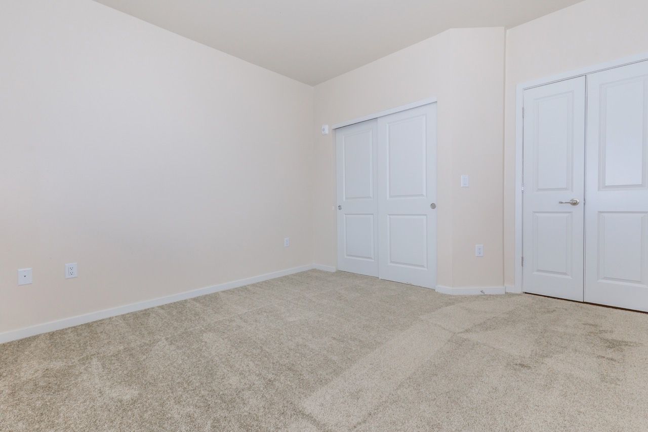 Empty beige bedroom with plush carpet, white closet doors, and an interior door.