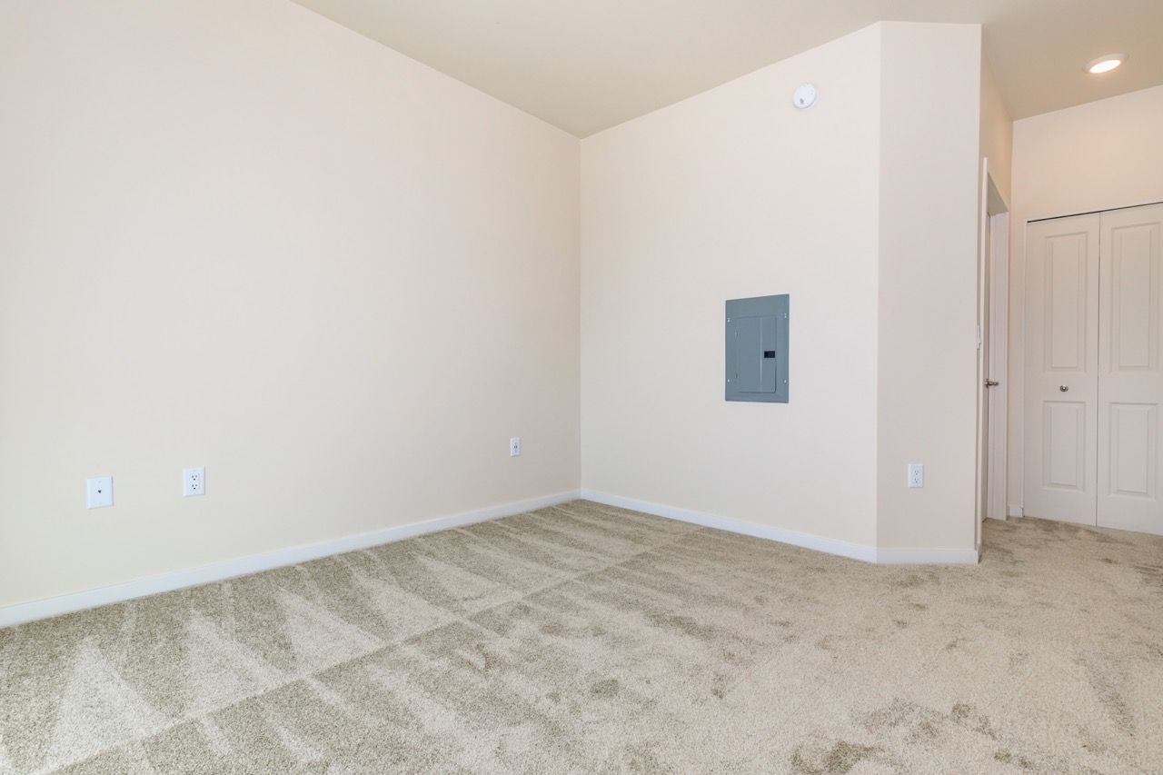 Empty beige apartment room with carpet, electrical panel on wall, and a doorway to the right.