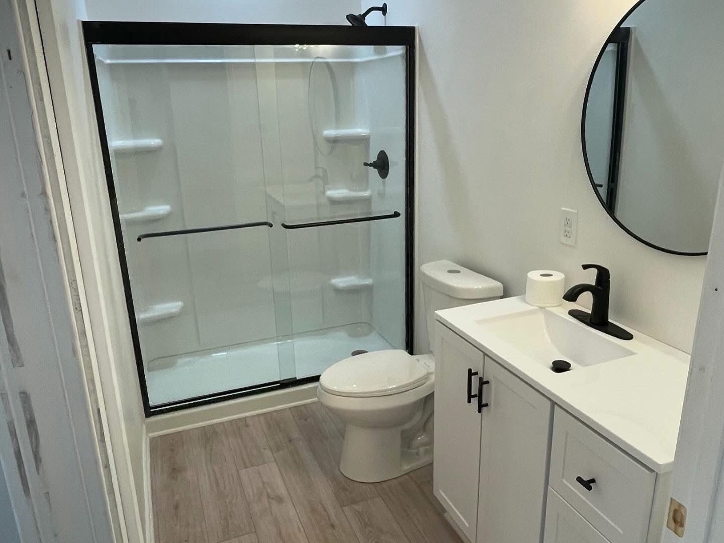 Small bathroom with white vanity, round mirror, toilet, and glass shower enclosure with wood-look floor.