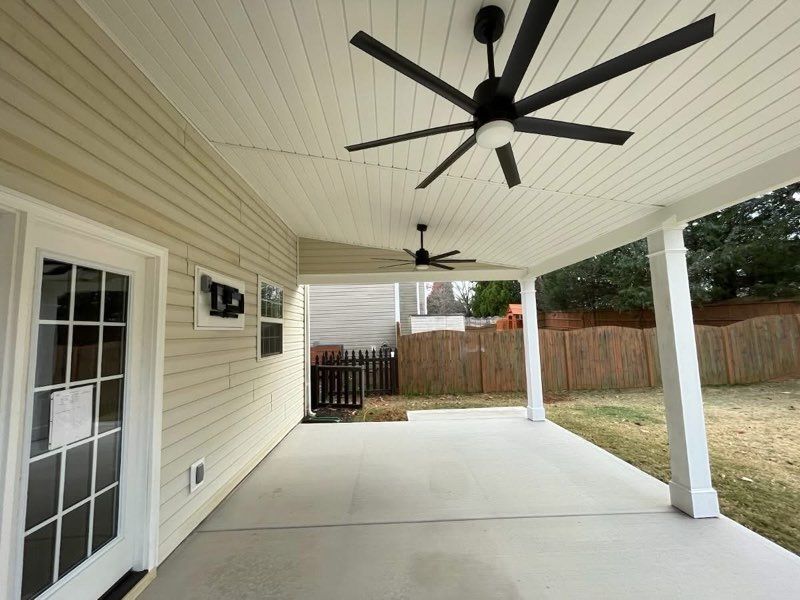 Covered concrete patio with white ceiling, black ceiling fans, and a screened door beside a fenced yard.
