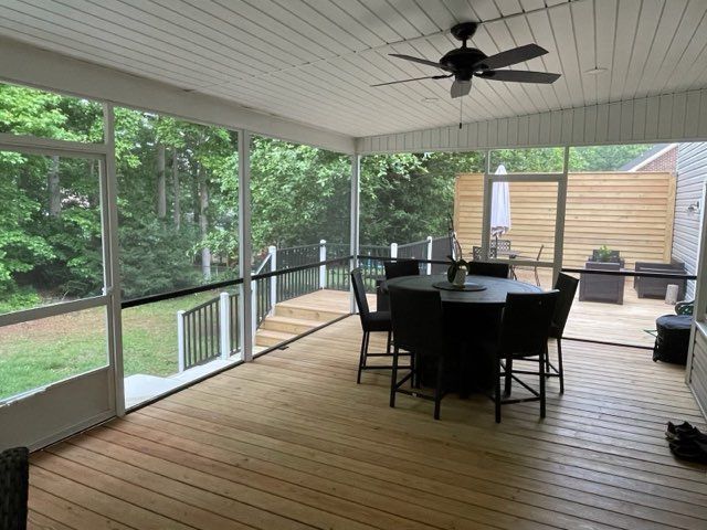 Covered wooden patio with dining table, chairs, ceiling fan, and view of a wooded backyard
