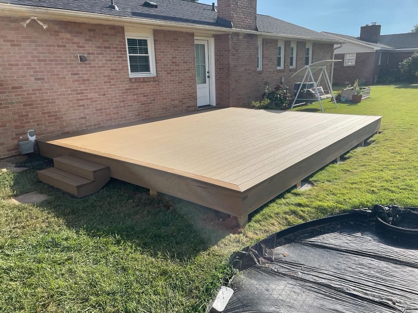 Raised wooden deck platform beside a brick house, with steps on the left and lawn around it