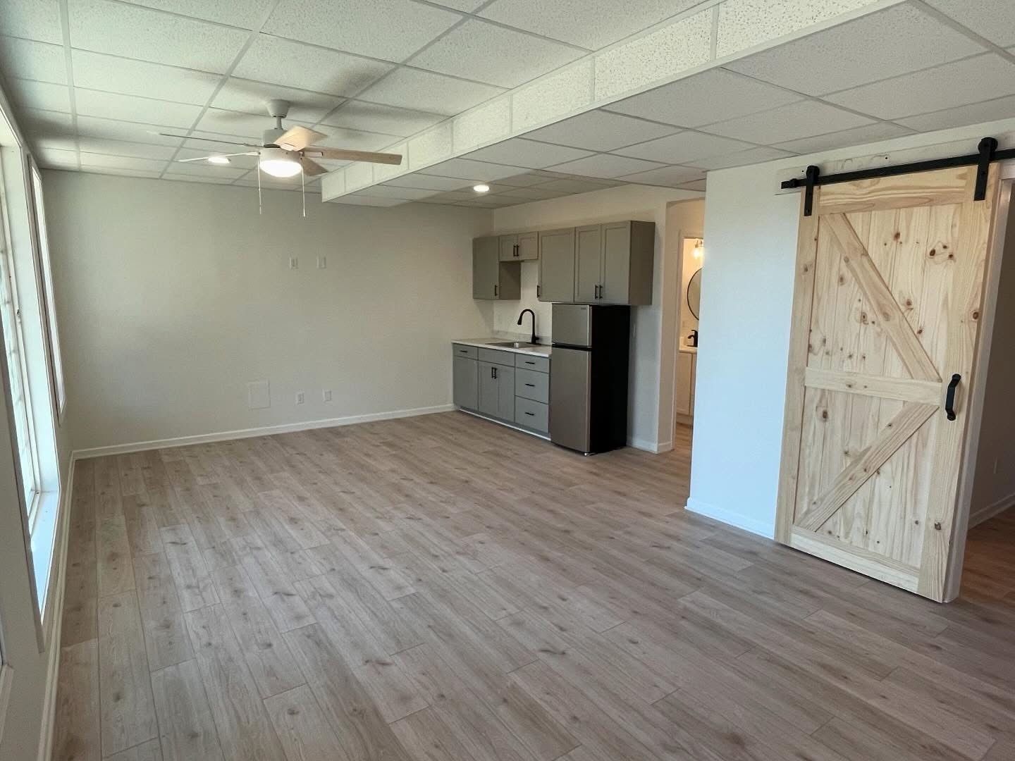 Empty room with wood flooring, ceiling fan, kitchenette, and a rustic sliding barn door