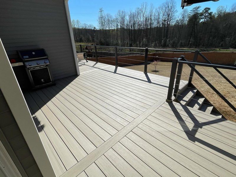 Sunny backyard deck with gray boards, railing, grill, and stairs leading to a yard and trees