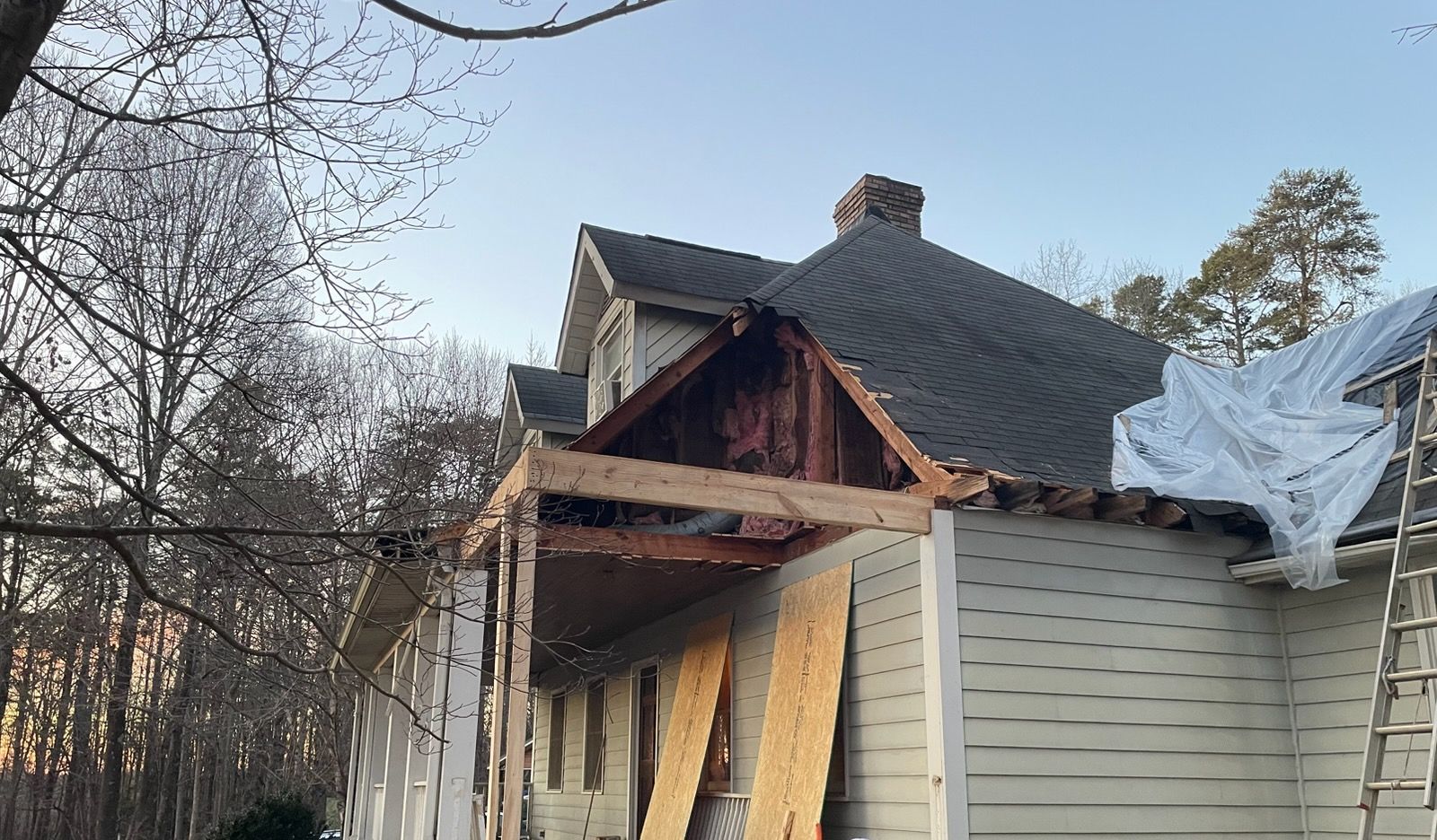 Partially demolished house with exposed roof framing and tarped damage near trees