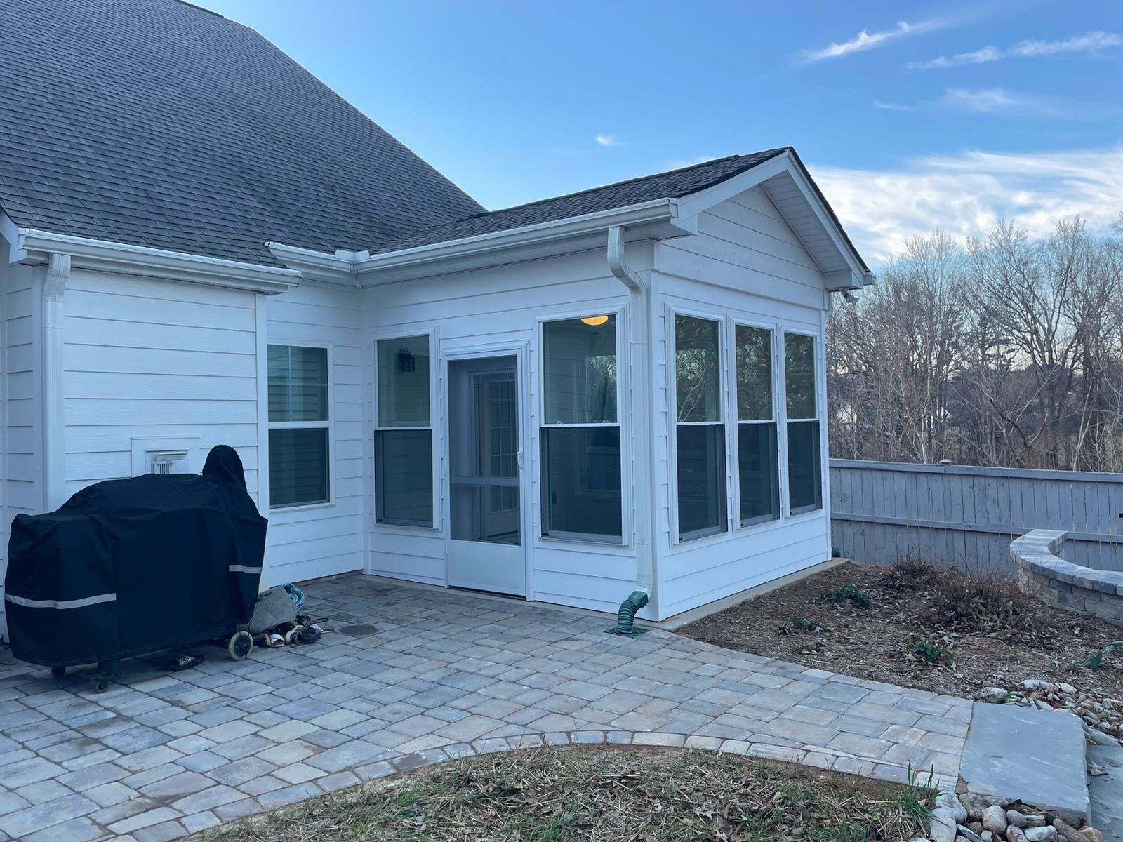 White house sunroom with many windows, black grill, and paved patio beside a fenced yard