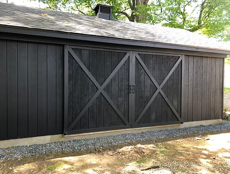 Dark gray wooden shed with double barn doors and X-bracing, set on gravel in a wooded yard.