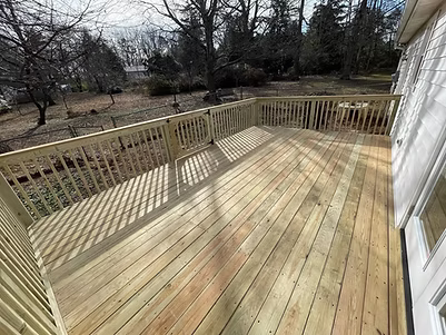 Wooden deck with beige railing beside a white house, overlooking a wooded yard