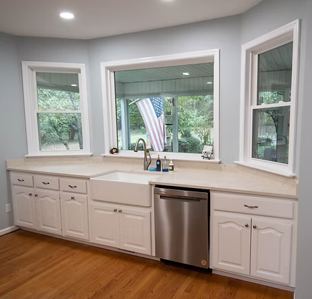 Bright kitchen with white cabinets, center sink, stainless dishwasher, and large windows overlooking greenery