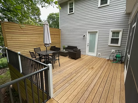 Wooden backyard deck with patio table, chairs, umbrella, and outdoor seating beside a gray house.