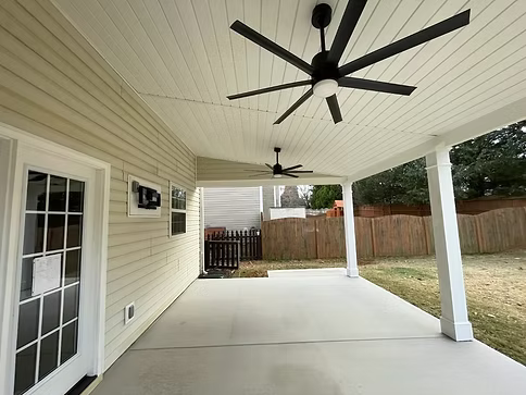 Covered patio with white ceiling, black ceiling fans, and a concrete floor beside a house and fenced yard