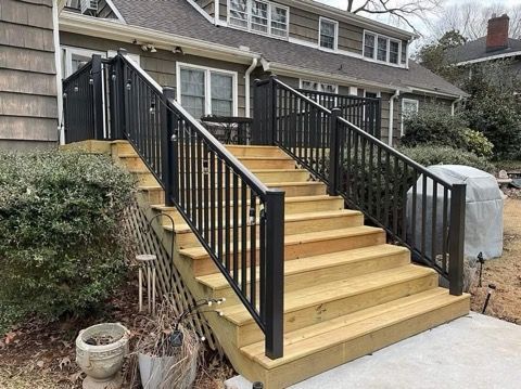 Front steps with black railings leading to a brick house entrance, with shrubs and a patio in front.