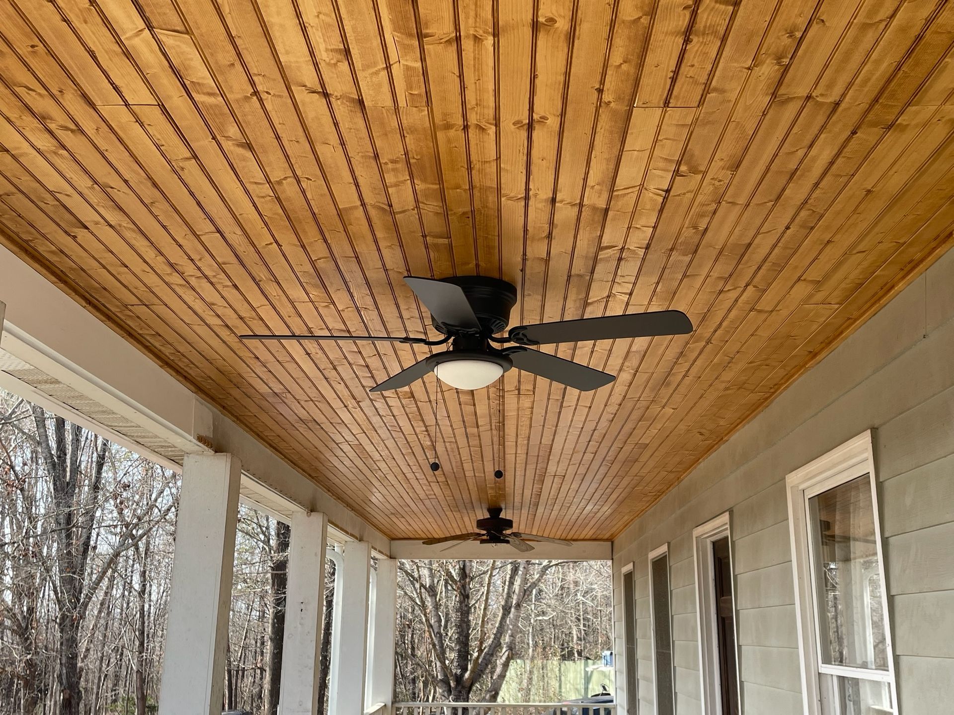 Covered porch with wood plank ceiling, ceiling fans, and screened windows overlooking bare trees.