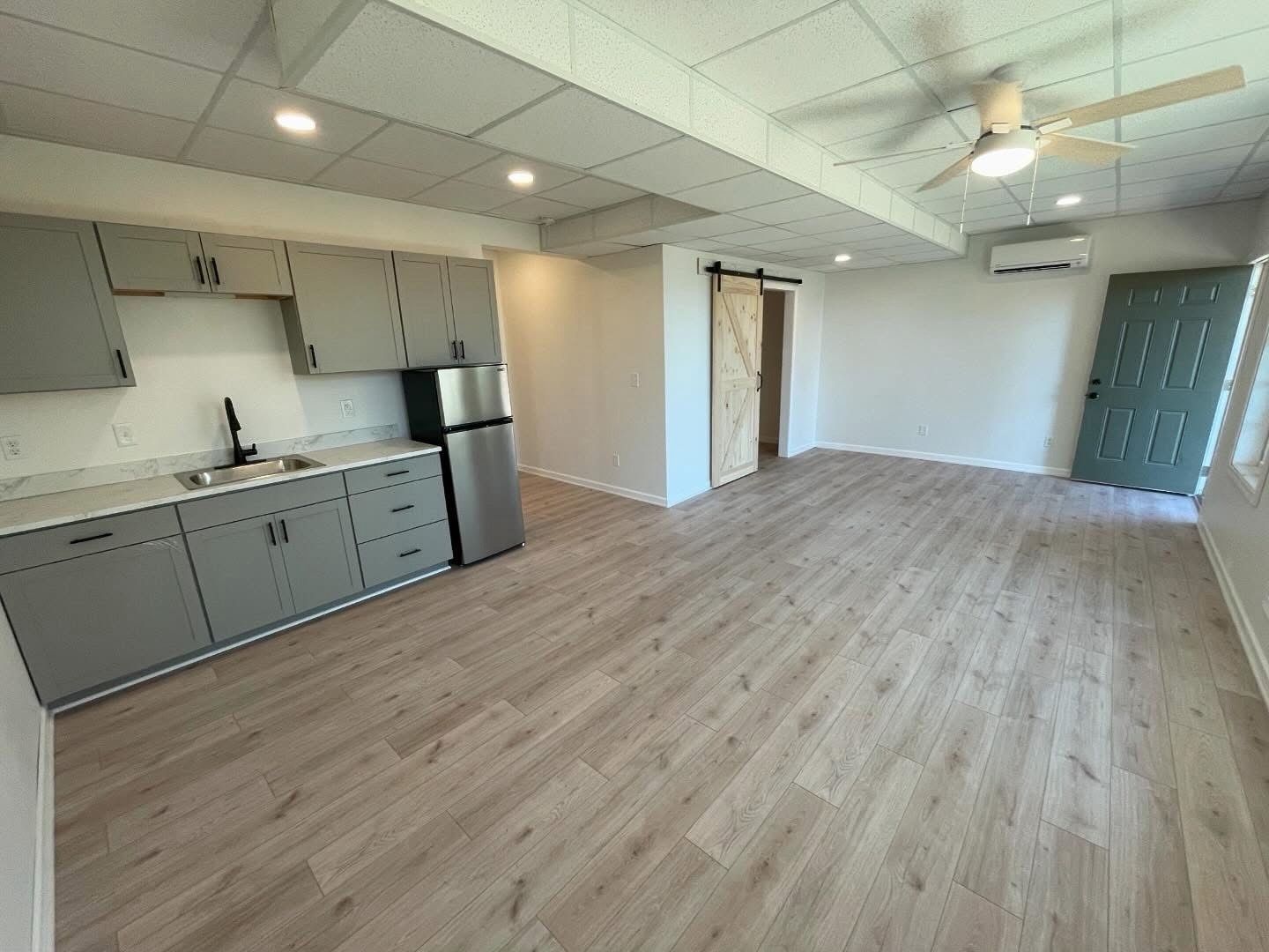 Empty open-plan studio with gray kitchen, wood floors, ceiling fan, and sliding glass door