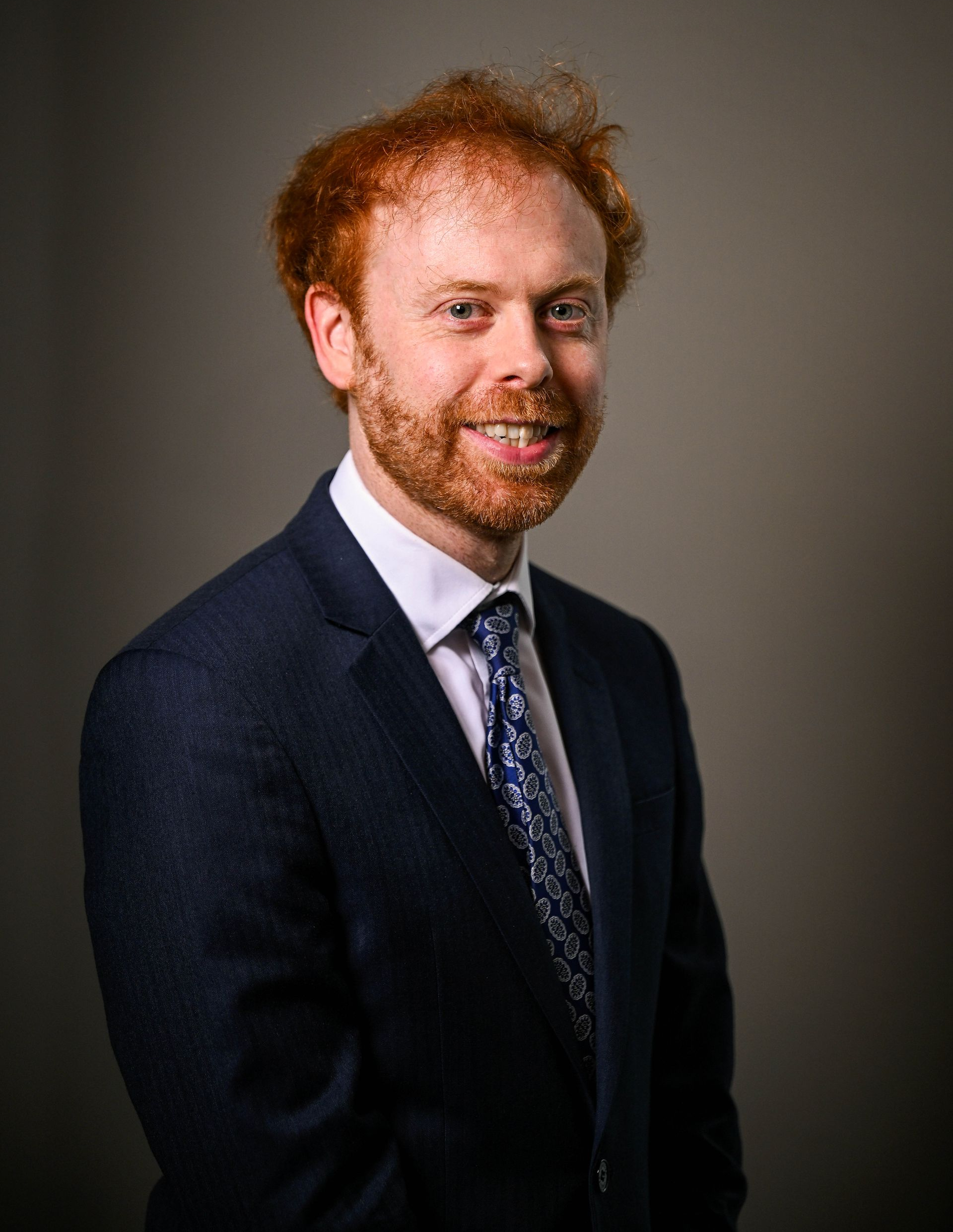 Man with red hair and beard in a suit, smiling, against a grey backdrop.