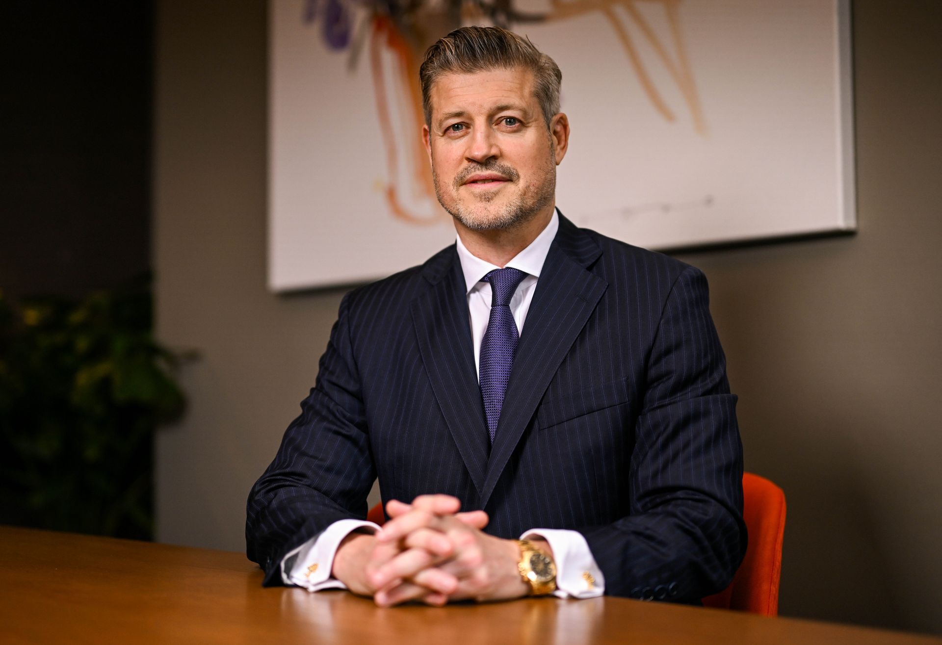 Man in suit, hands clasped on table, smiling slightly. Office setting, artwork on wall.