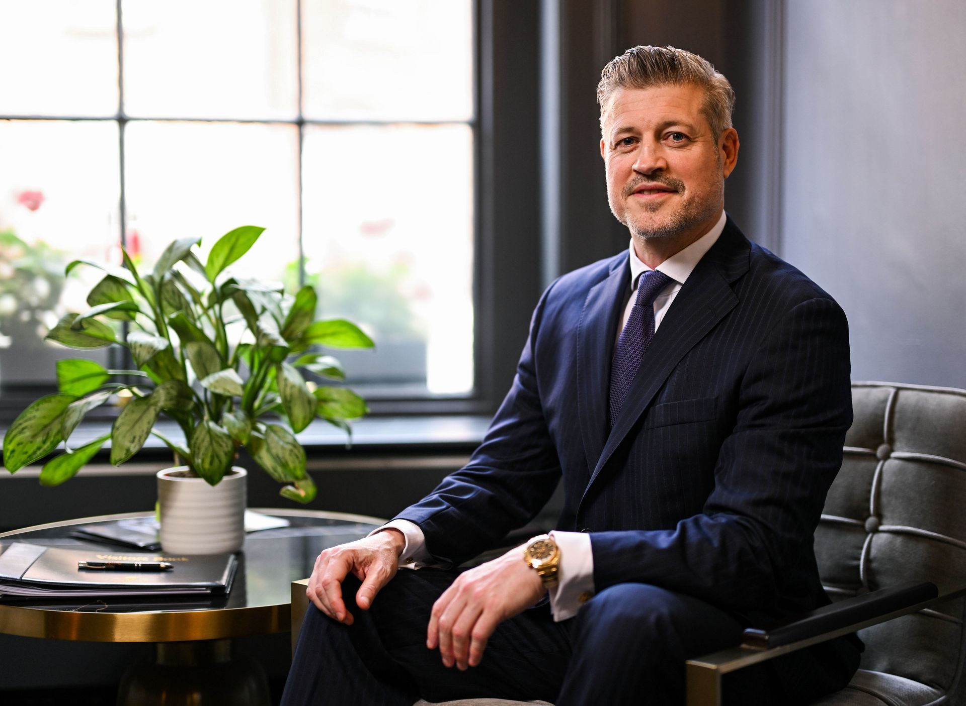 Man in suit seated in front of a window, smiling. A small plant is next to him.