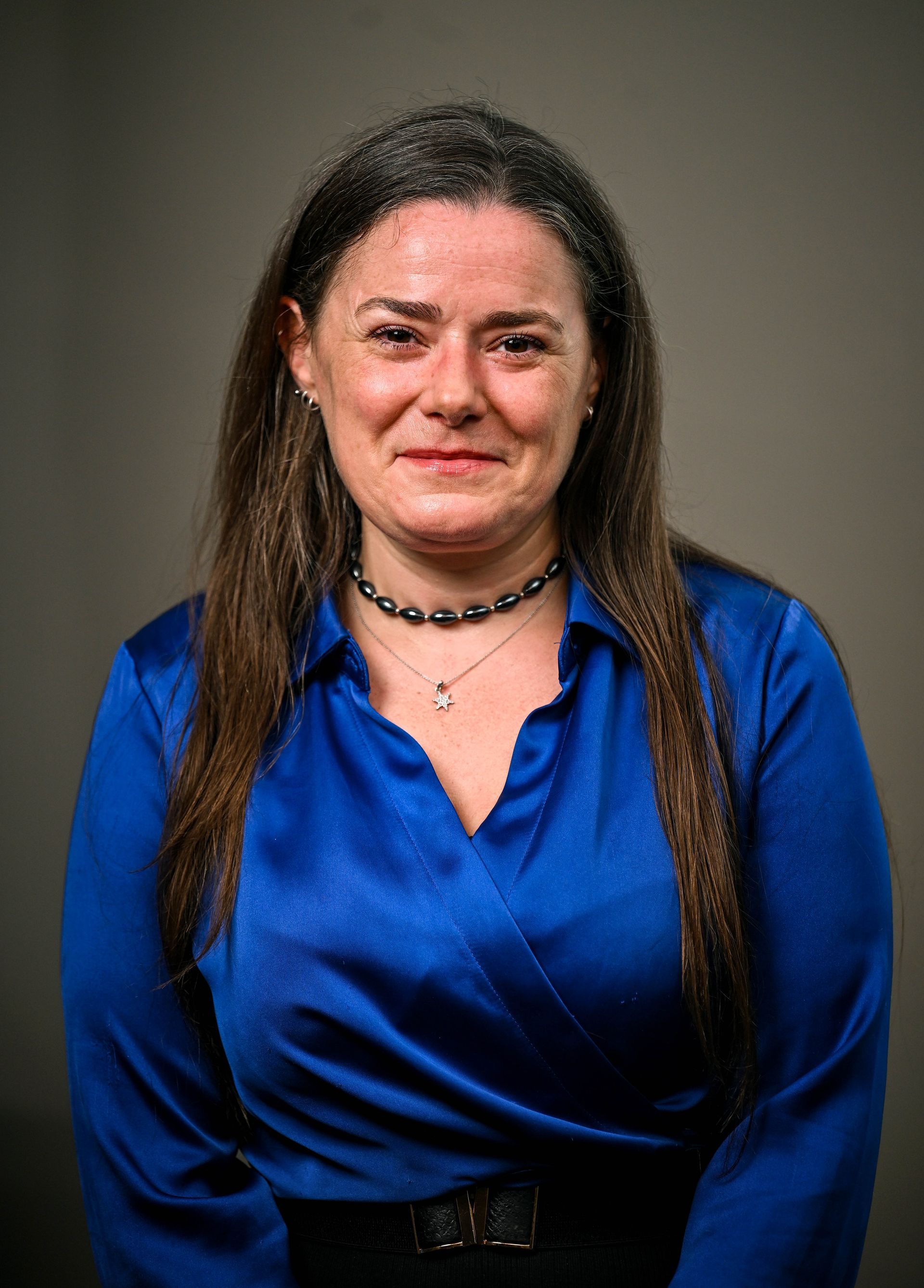Woman with brown hair, blue shirt, pearl necklace, smiles, gray background.