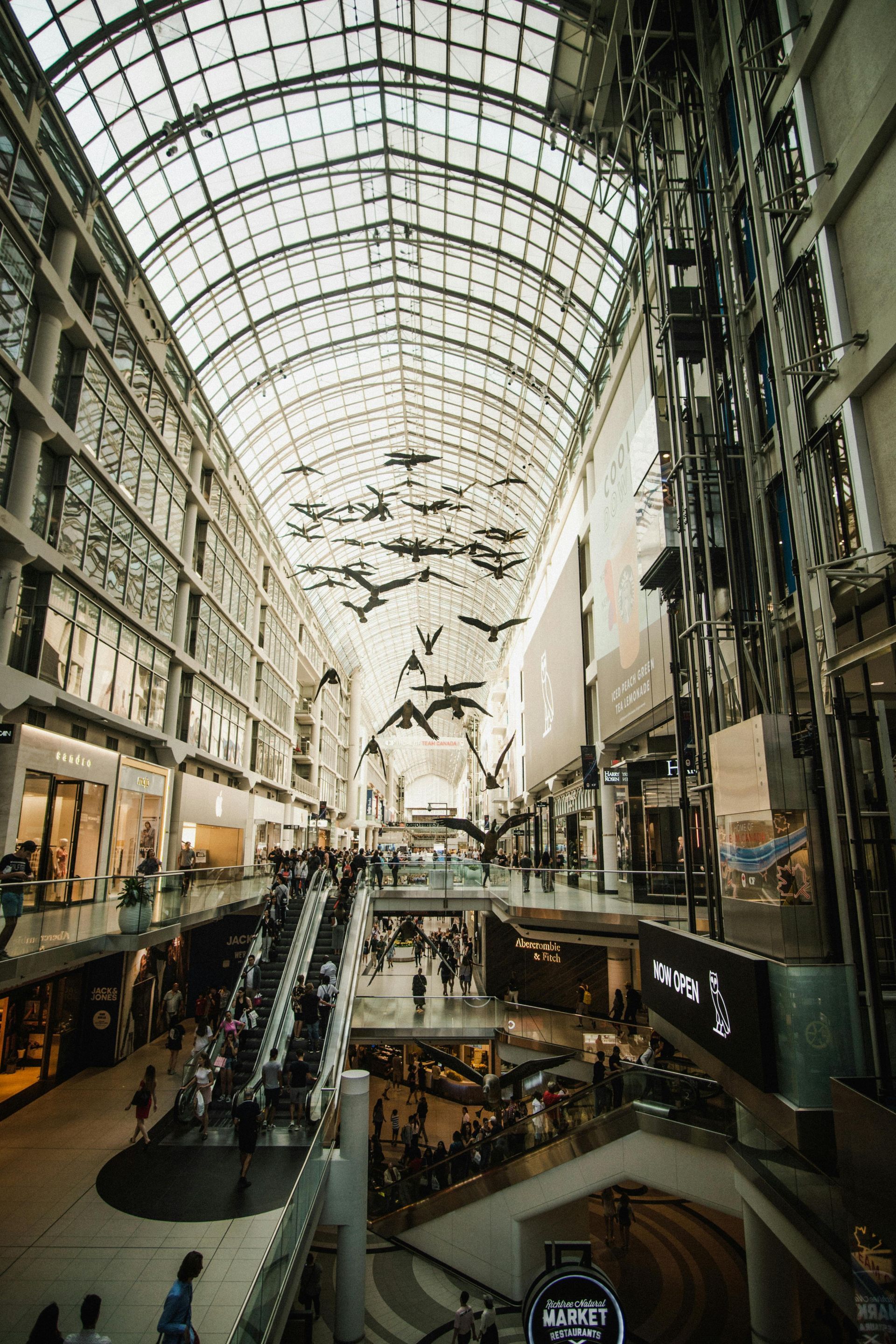 Interior of a shopping mall with escalators, glass ceiling, and shoppers, a sculpture of flying birds.