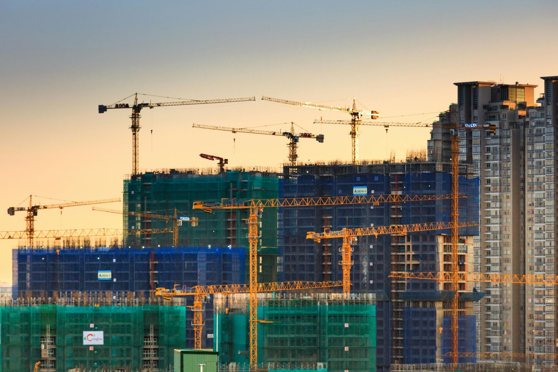 Construction site with numerous cranes and partially built high-rise buildings, blue and green netting, golden hour lighting.
