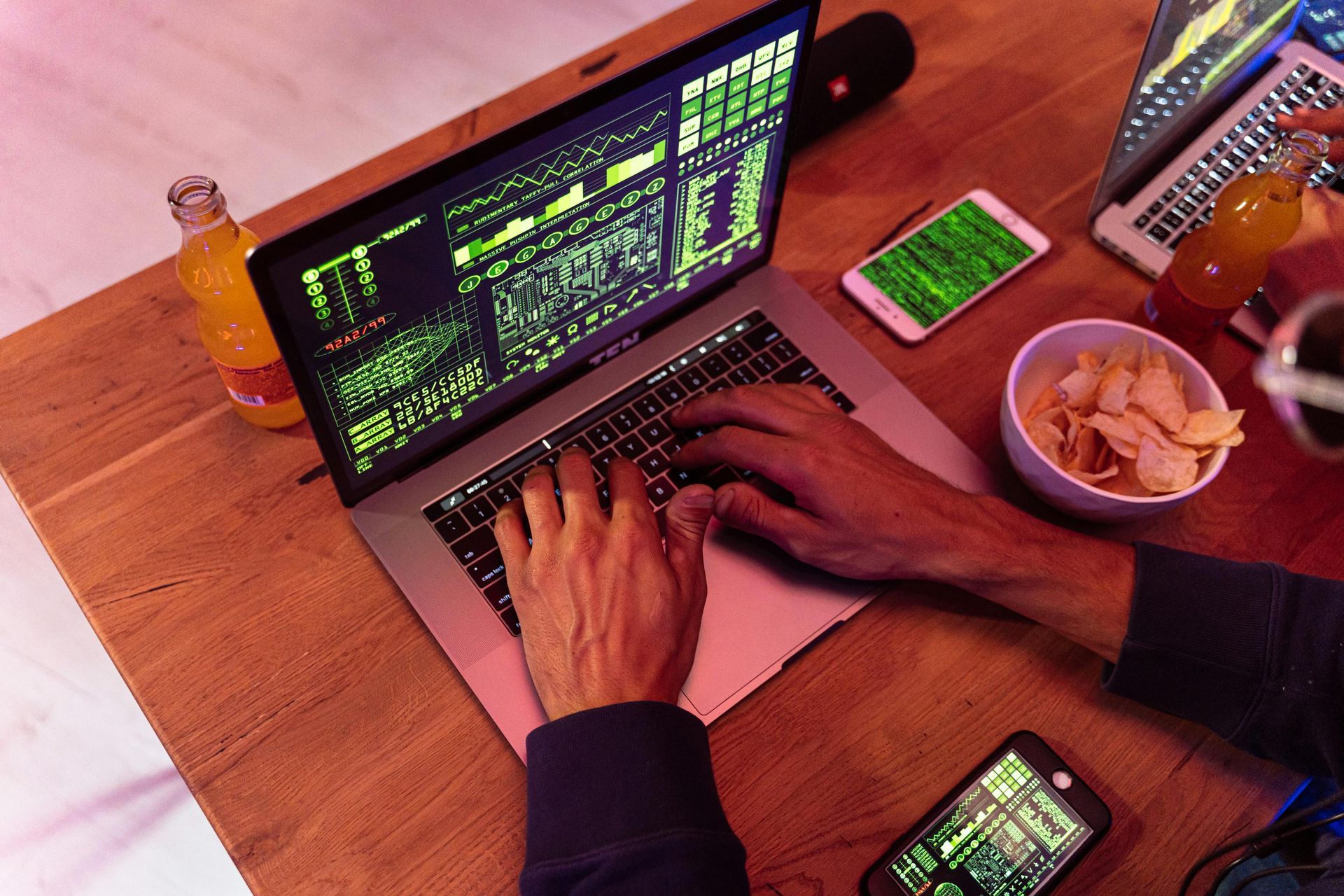 Person typing on laptop with green code, surrounded by phones, snacks, and drinks.