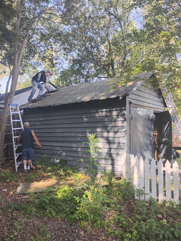 People working on the roof of a small gray shed beside a white picket fence and ladder.