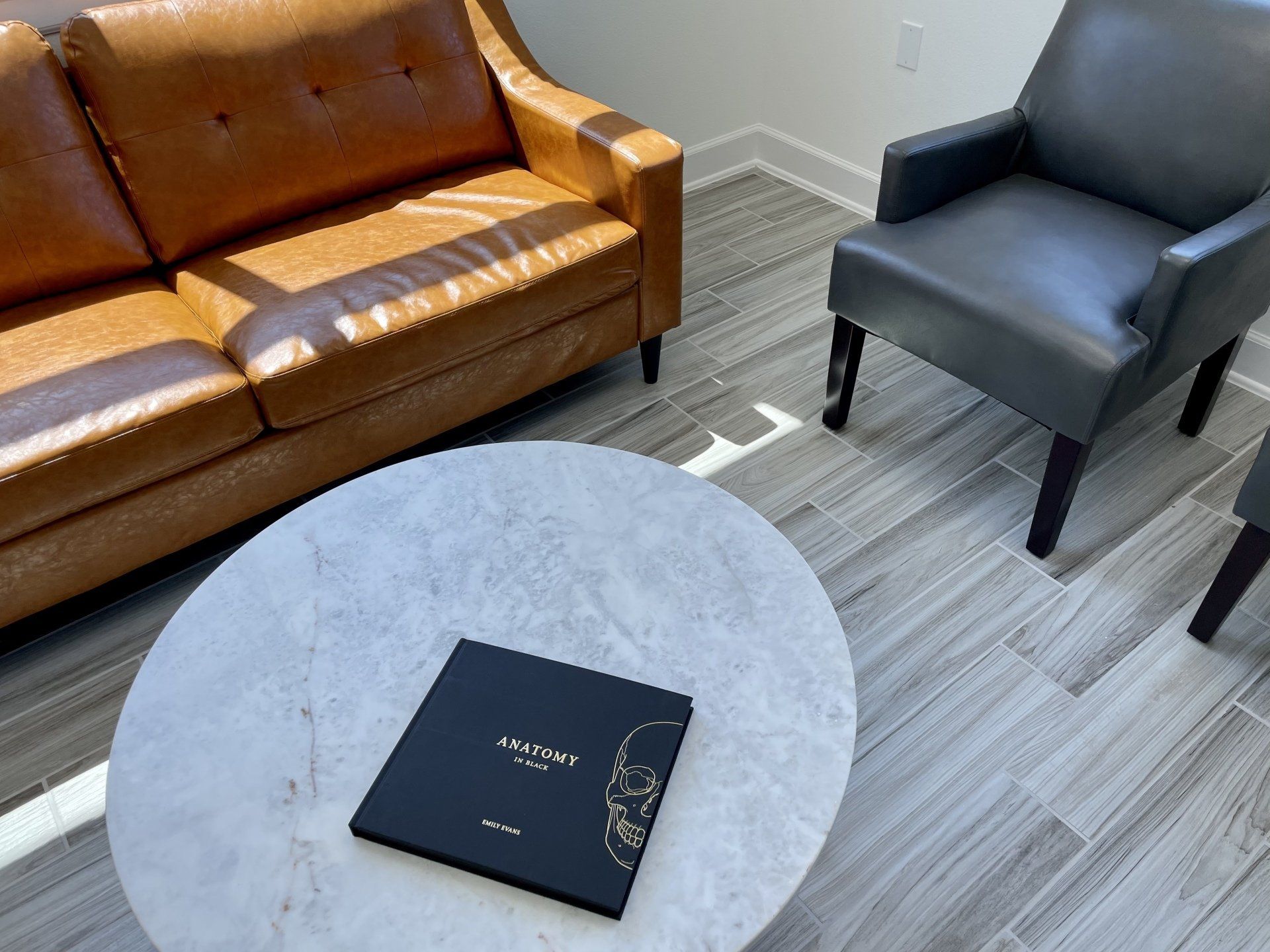 Waiting room with leather sofa, gray chair, round marble table and an anatomy book.