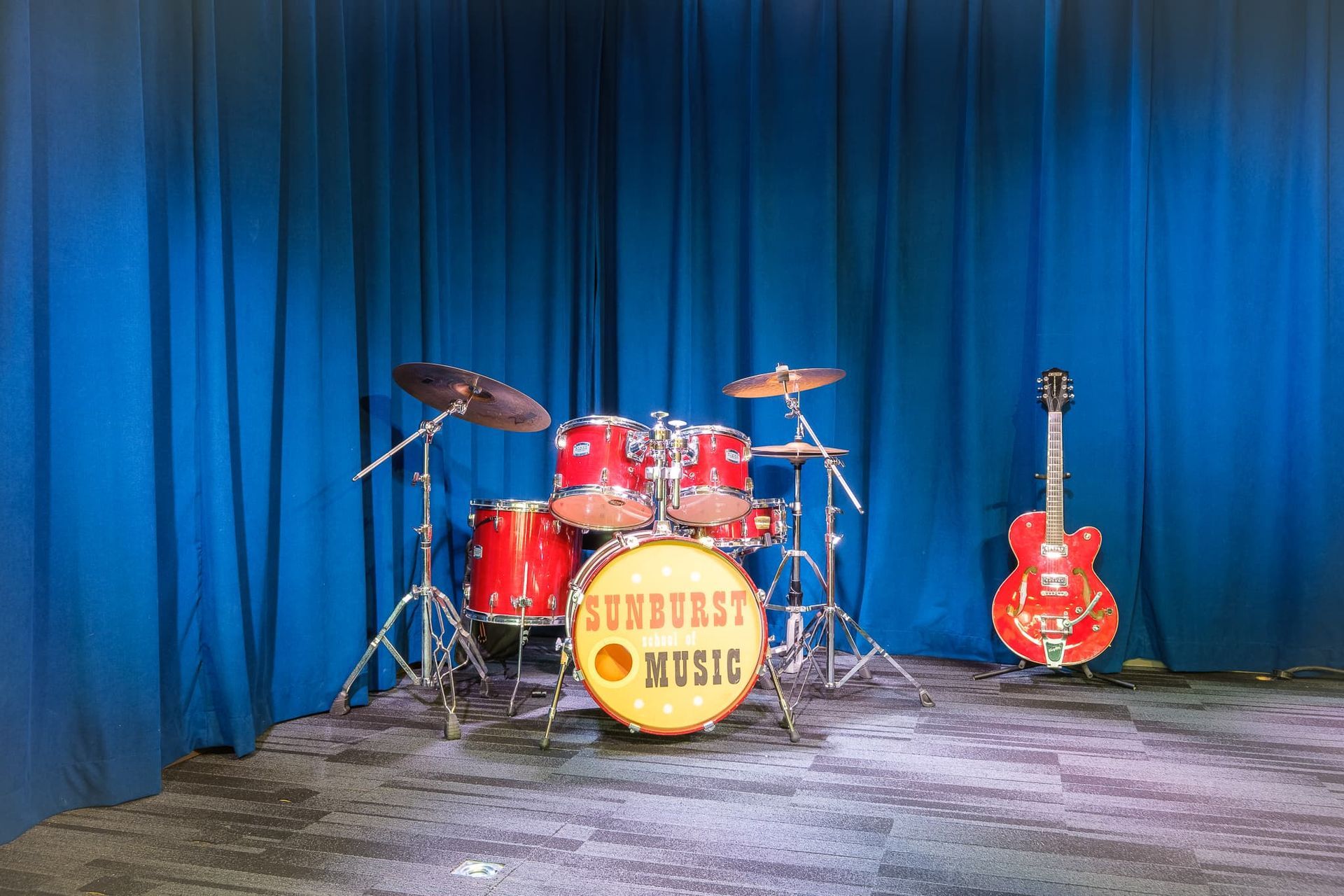 A drum set and a guitar are on a stage in front of a blue curtain.