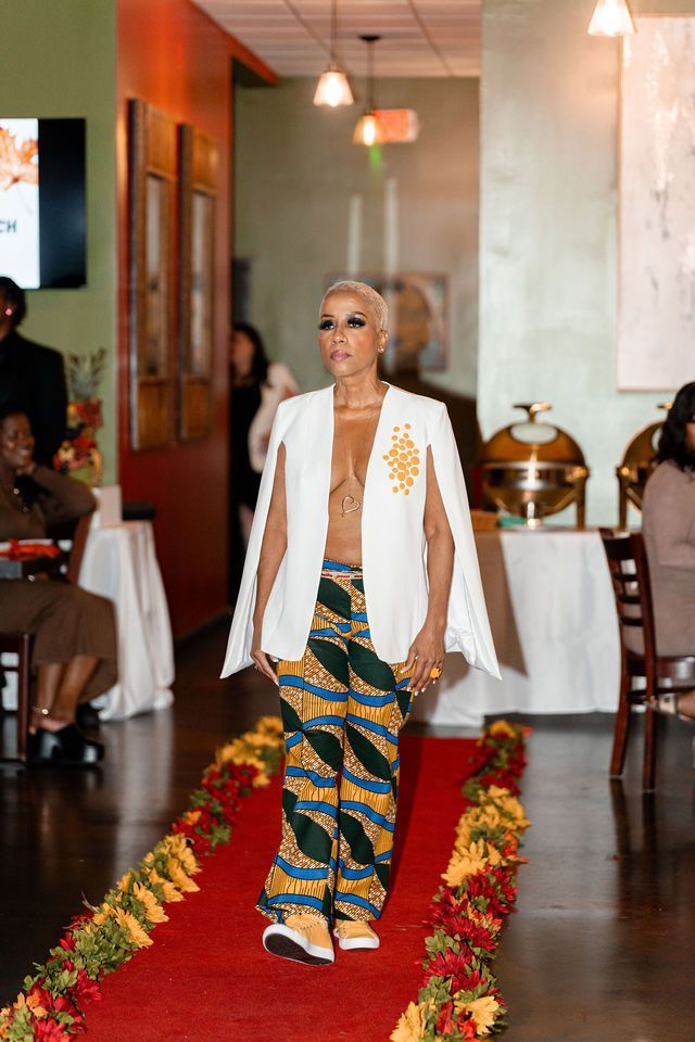 Woman modeling on a runway in white jacket, patterned pants, and sneakers. Red carpet, restaurant setting.