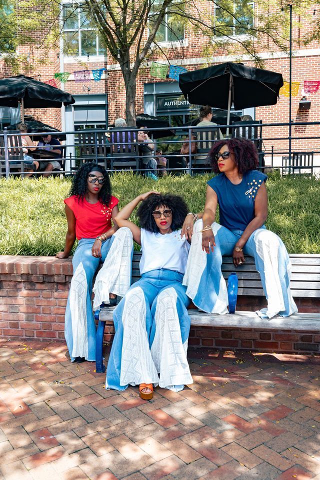 Three women sitting on a bench; wearing colorful tops and bell-bottom jeans, sunny outdoor setting.