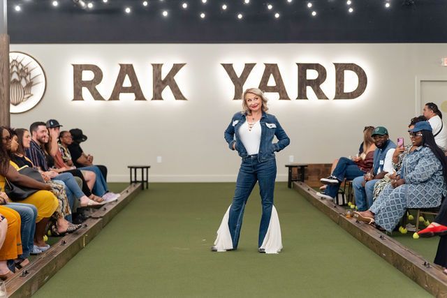 Fashion show at Rak Yard. Model in denim outfit poses on green runway as audience watches.