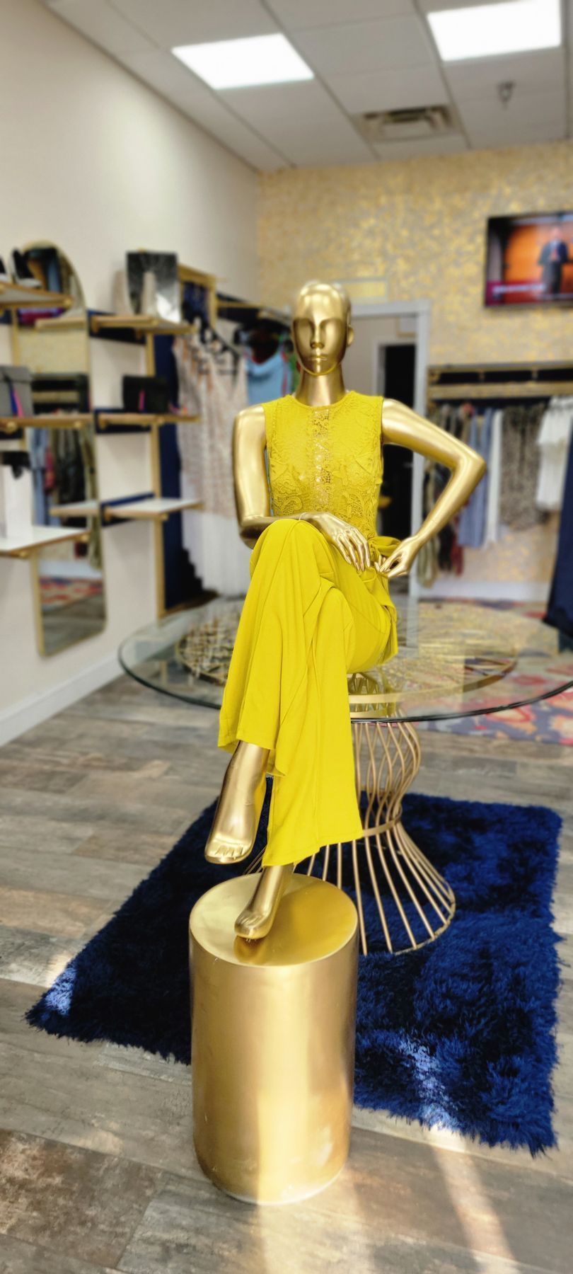 A gold mannequin wearing a yellow outfit sits on a glass table in a clothing store, with a gold stool.