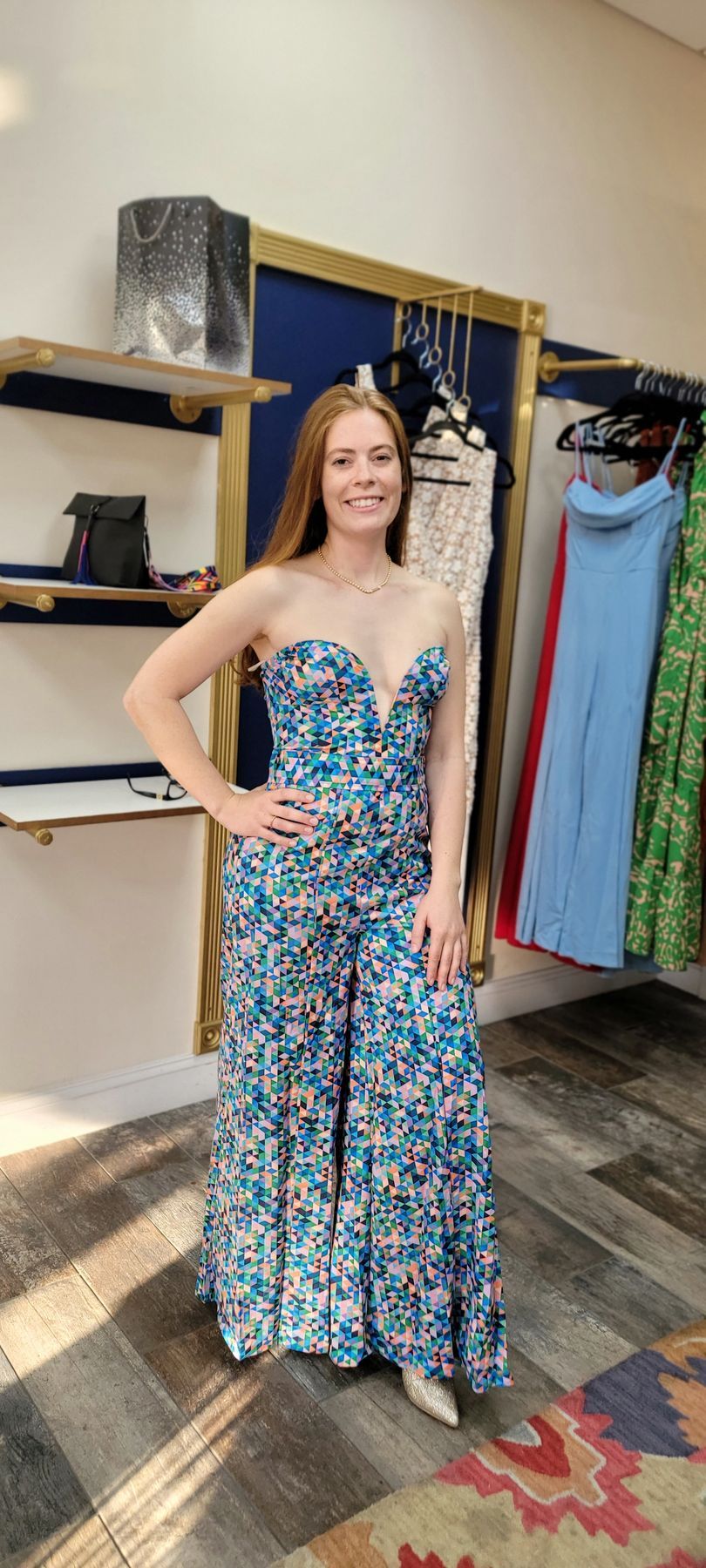Woman in floral jumpsuit smiles in a clothing store. She stands on a patterned rug near racks of clothes.