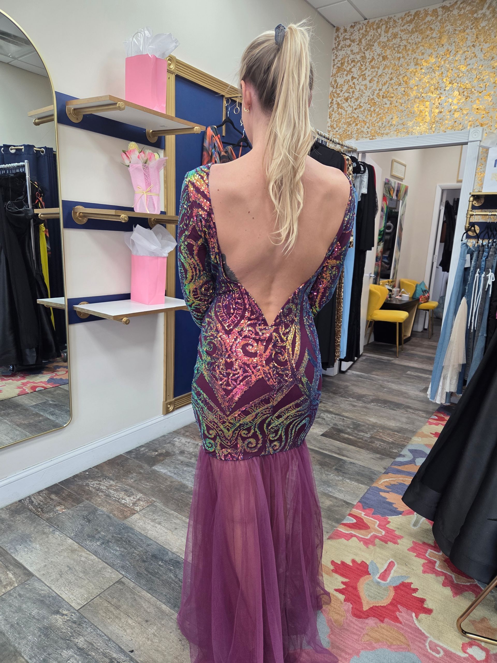 Woman in sequined gown with open back, standing in a boutique with dresses on racks.