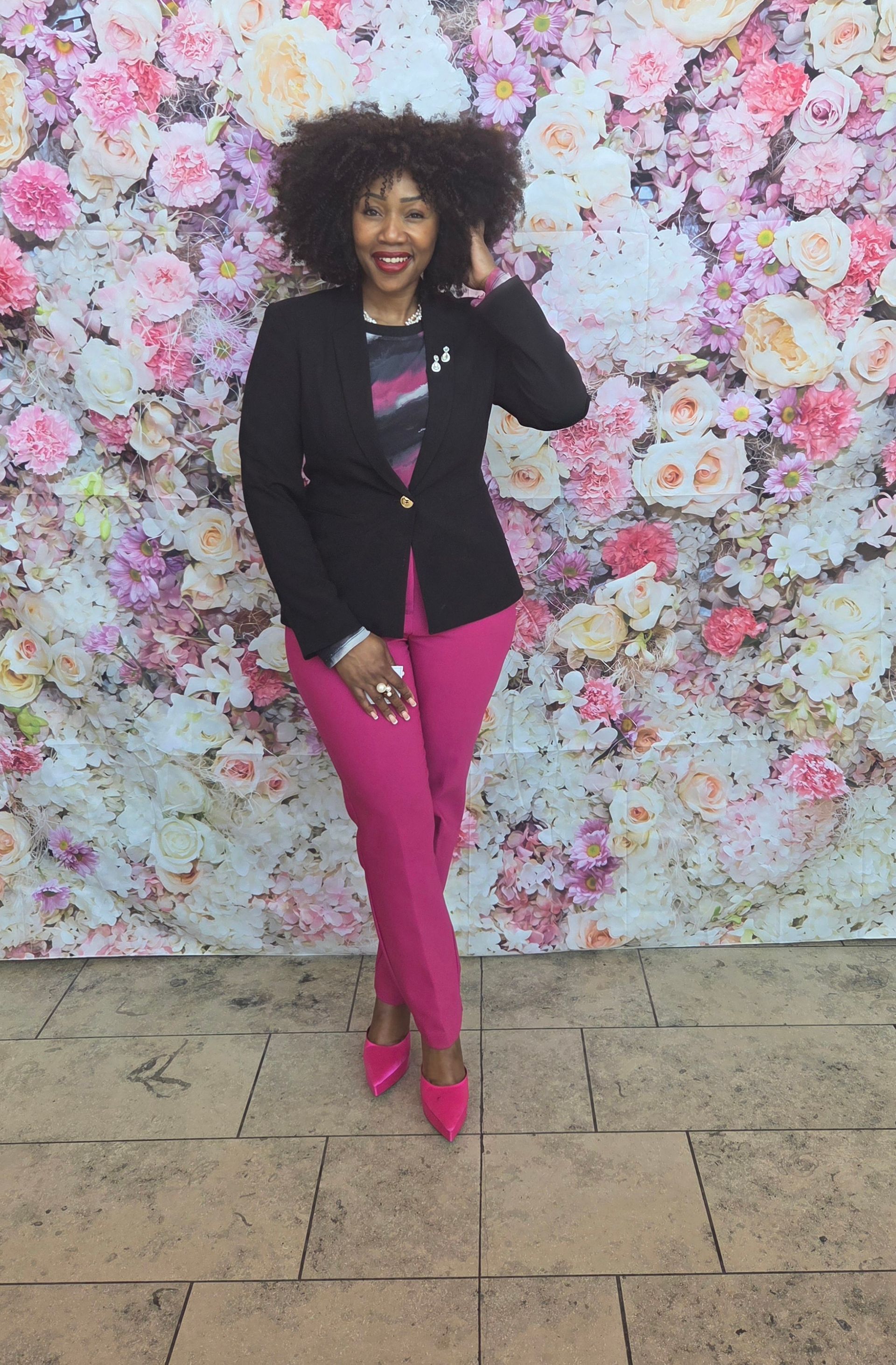 Woman in black blazer and pink pants poses in front of floral wall.