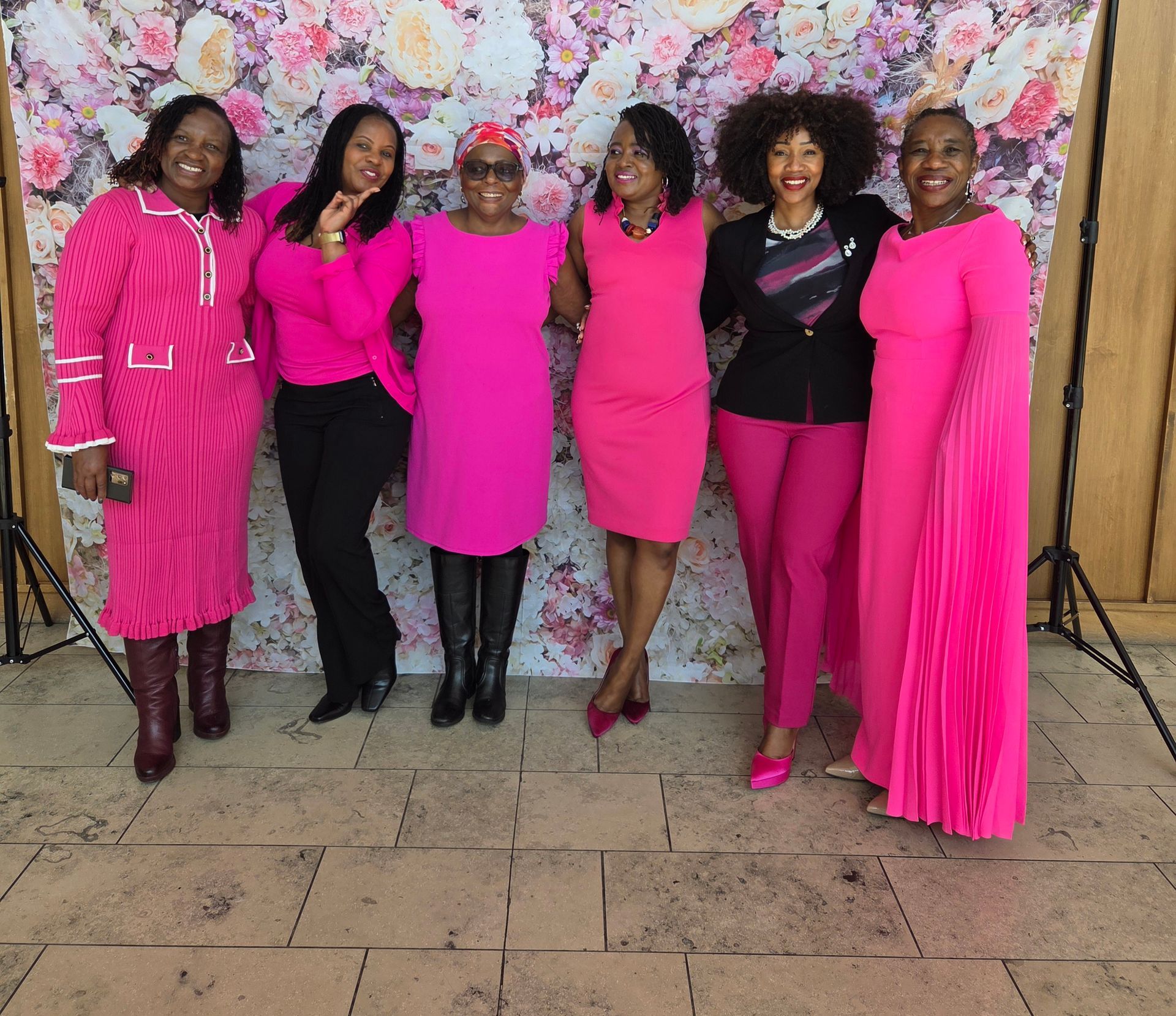 Six women in pink outfits pose in front of a floral backdrop.