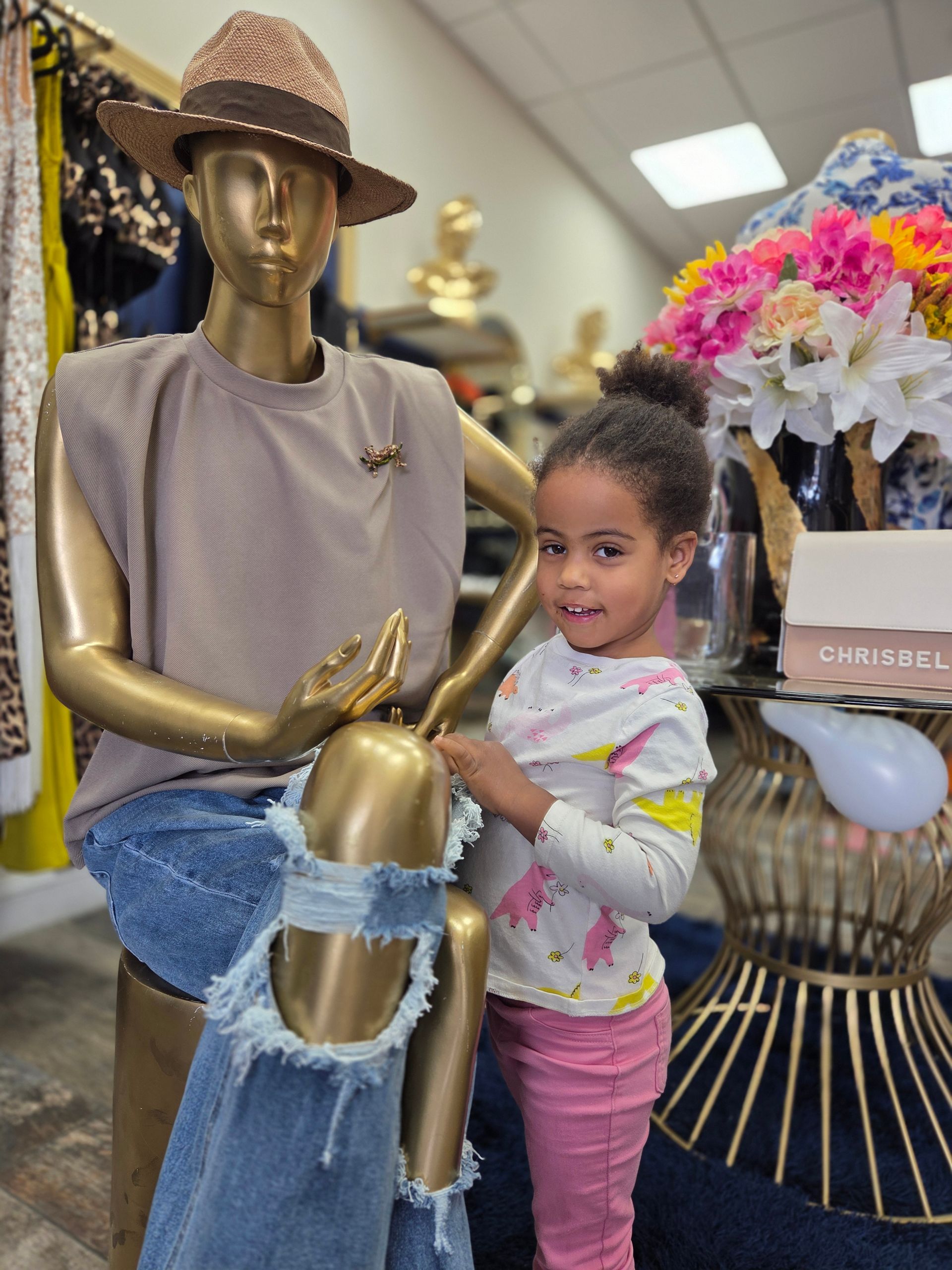 A young child smiles next to a gold mannequin wearing a denim dress in a clothing store.