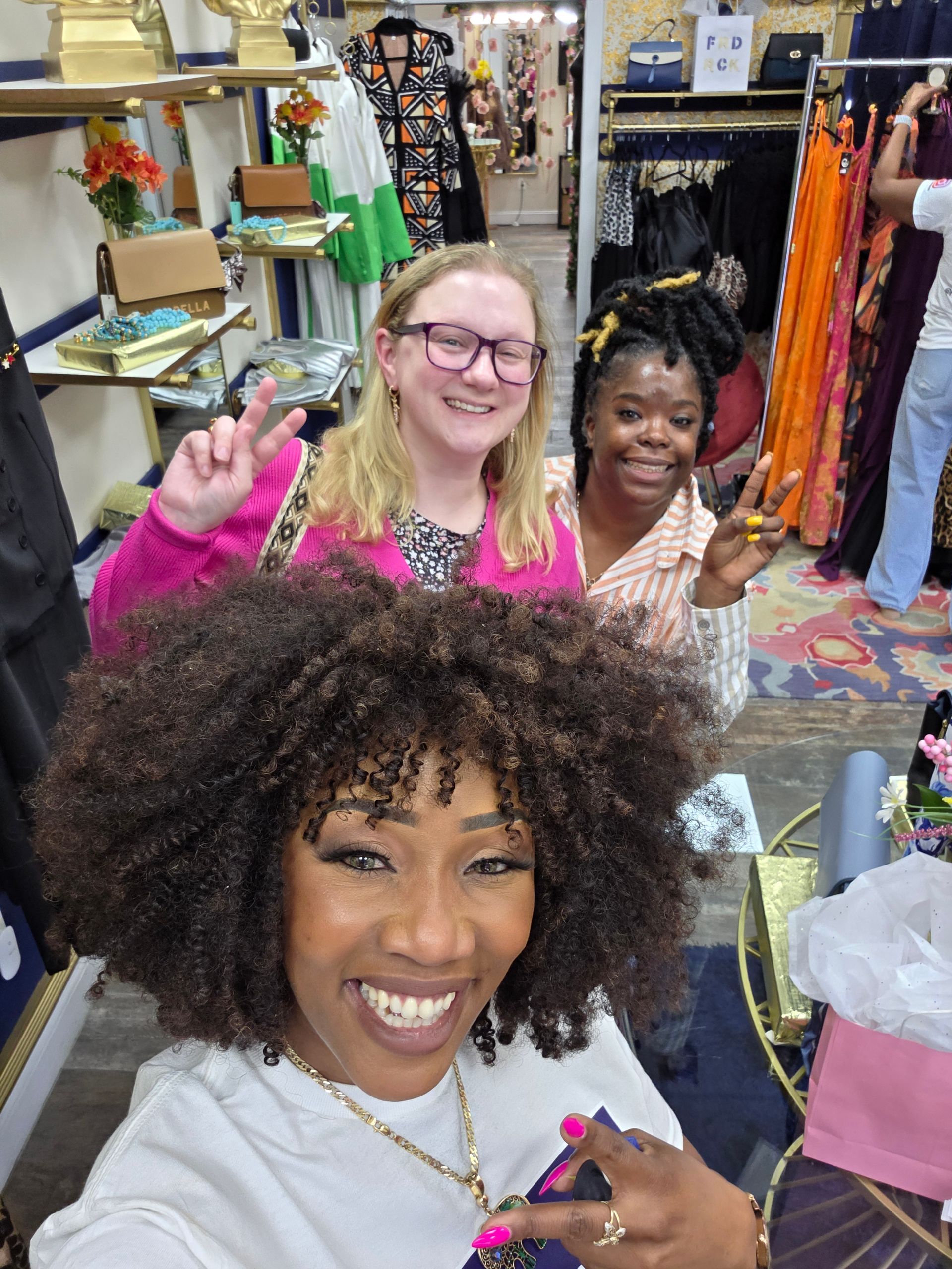 Three smiling people in a shop, making peace signs. One takes selfie. Shop has clothes and decor.