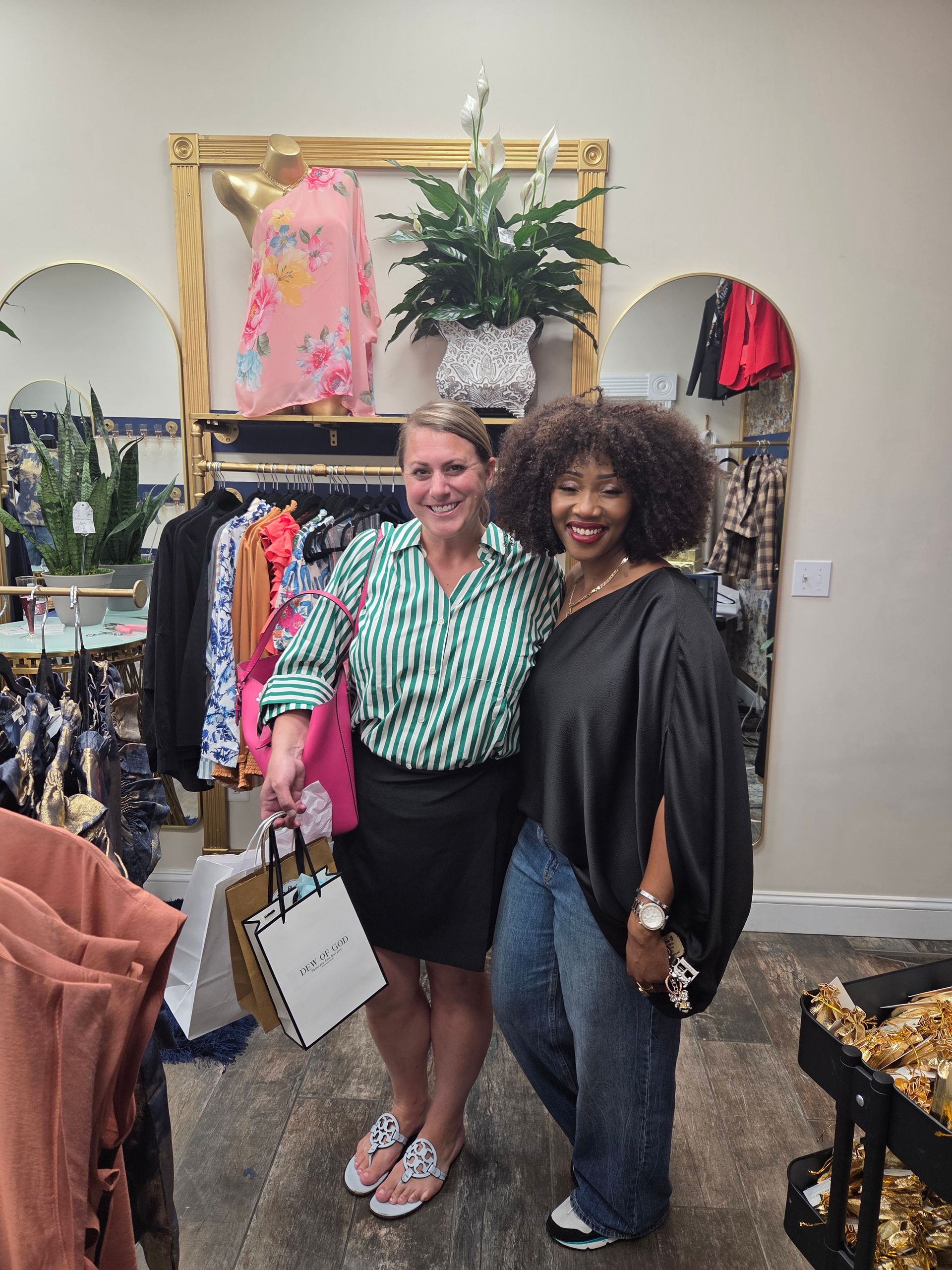 Two women smiling inside a clothing store. One holds shopping bags, the other wears a black top and jeans.