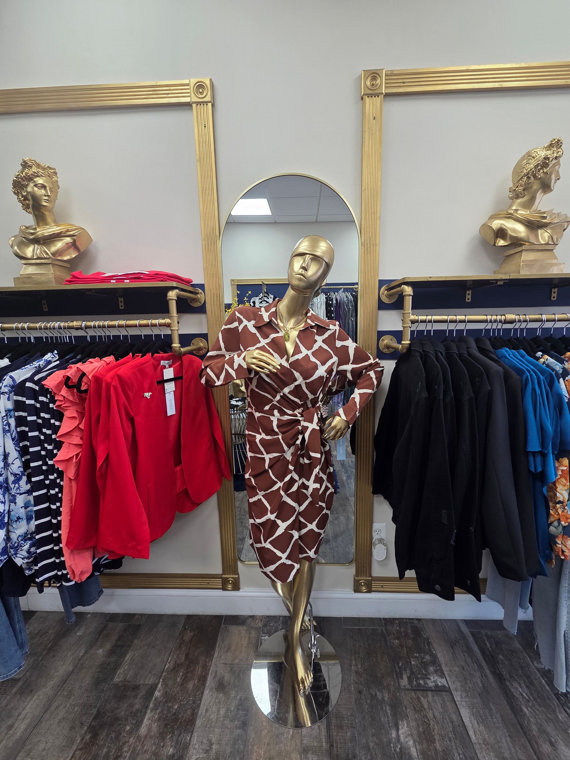 Gold mannequin in brown patterned dress, stands before a mirror, clothing racks on either side.