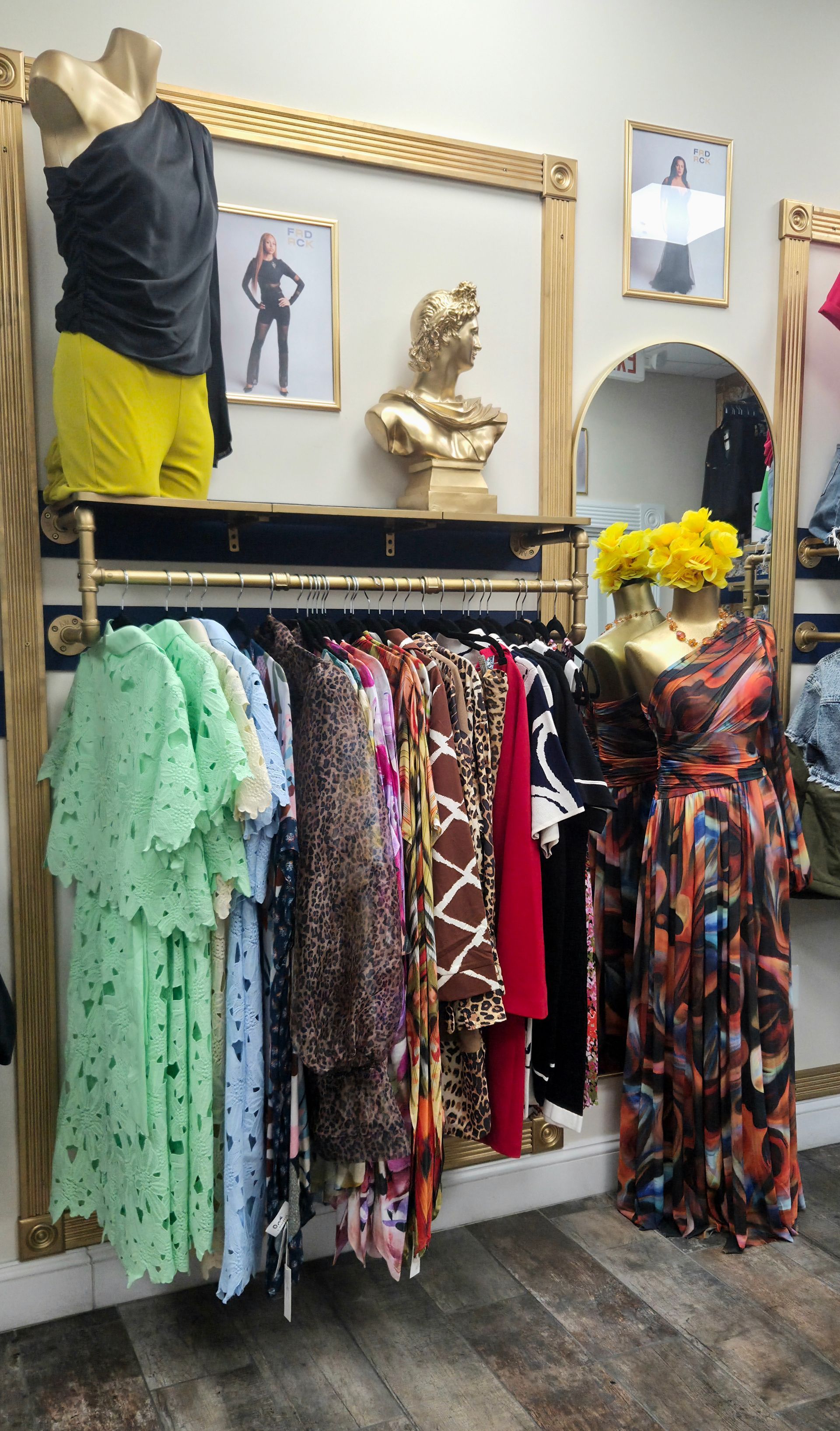 Clothing rack in boutique, featuring dresses and blouses, gold accents. Mannequins wearing yellow and floral designs.