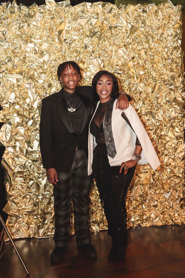 Two people pose in front of a gold wall. One person wears a black suit, the other a black outfit with a white jacket.
