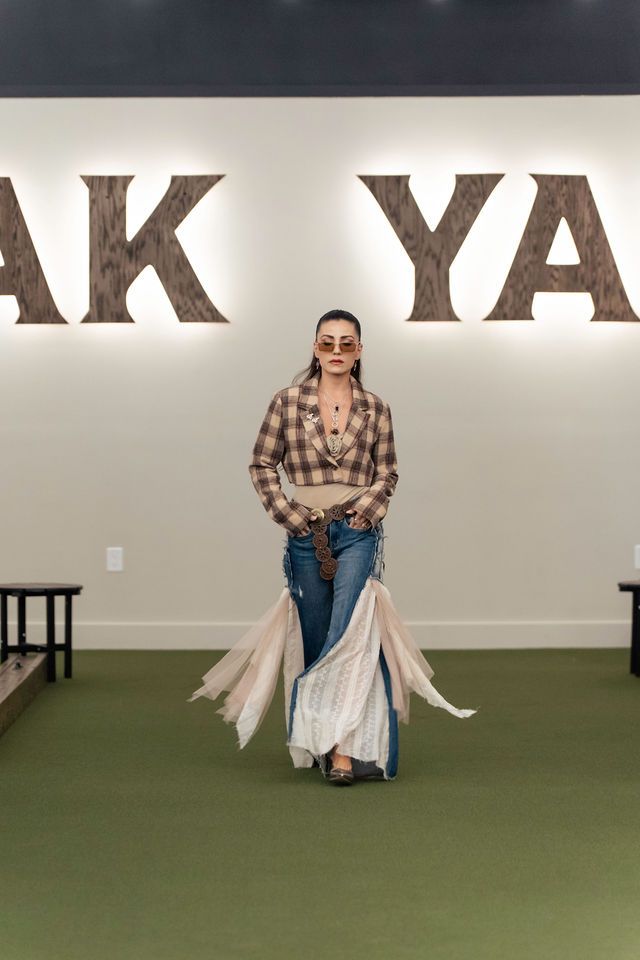 Woman in plaid top, denim skirt with flowing fabric, walking in front of a “YAK YARD” sign.